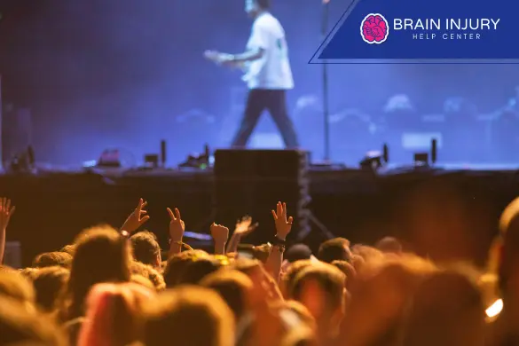 Multitud de concierto enérgica con manos levantadas, escenario con guitarrista y logo de Brain Injury Help Center.